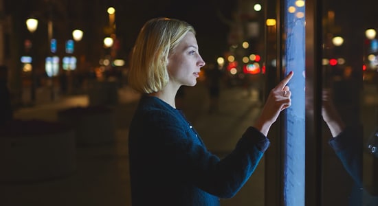 Young caucasian woman using a touch sensitive display while standing on the street with night lights on background, female touching futuristic smart bus station display for self service information
