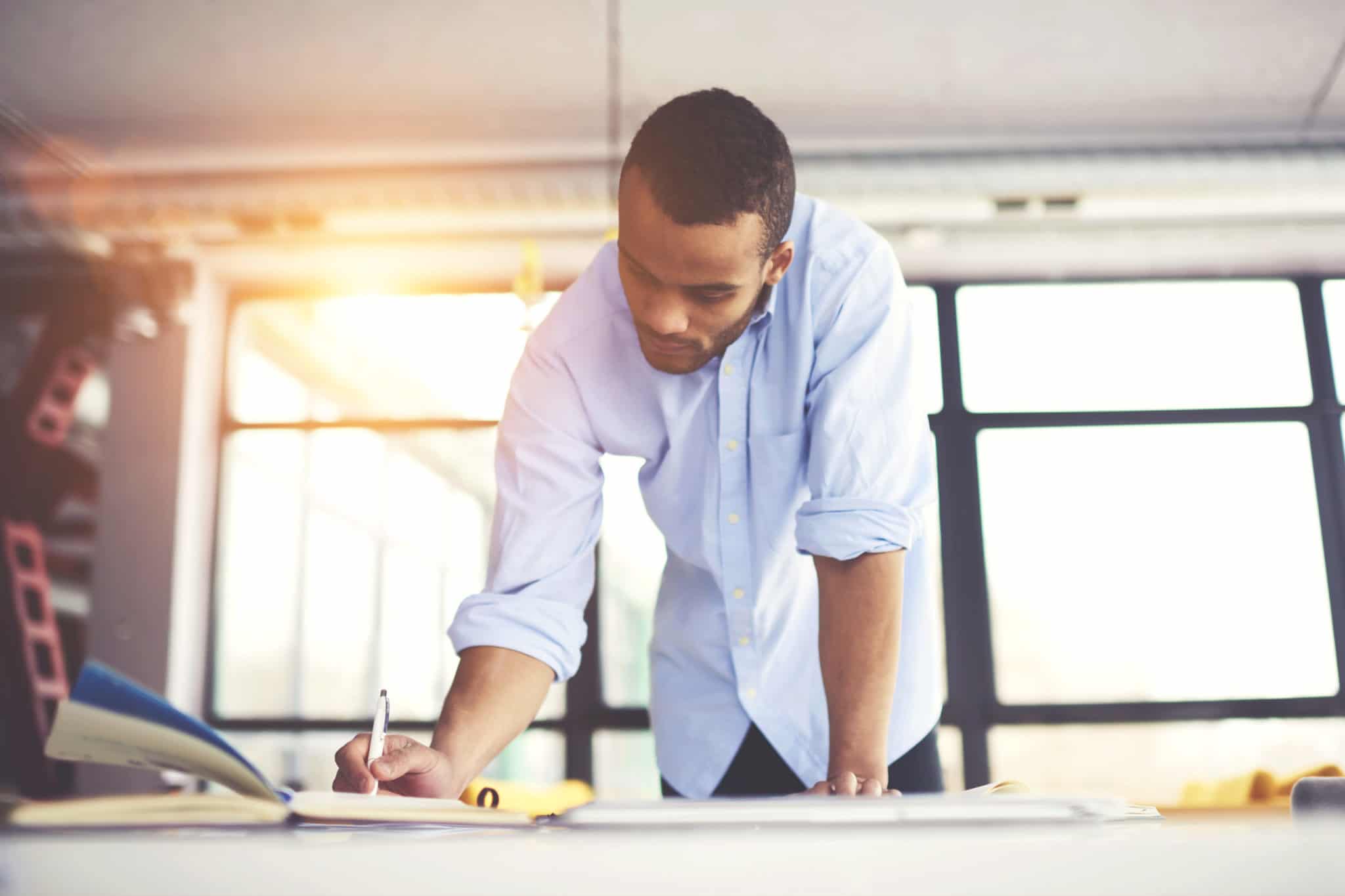 Skilled male afro american engineer thinking over blueprint improvement noting ideas in copy book to correct mistakes in planning looking concentrated on workspace desk with papers in modern studio