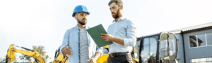 Male engineering manager in a suit showing another male engineer wearing a hard hat a clipboard with instructions whilst on a construction site