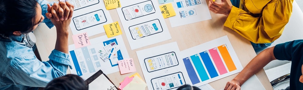 A group of creatives are sat around a table. On the table are documents that relate to different parts of the design process, along with images of mobile phones as they are working out the user interface.