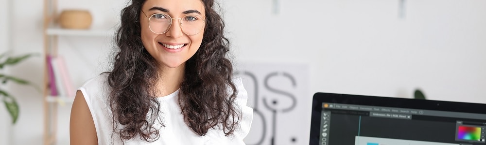 Young female digital design and communication manager with long brown hair, gold round glasses and a white dress stood up smiling at the camera in a small workspace stood infront of a computer displaying digital design software