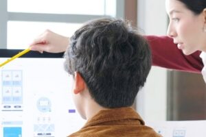 Female Digital Design Manager with a black ponytail, white shirt and maroon jumper answering the question ‘What is human centred design?’ by pointing to a desktop computer screen with a pencil explaining the concept to a male Digital Designer wearing a light brown jacket.