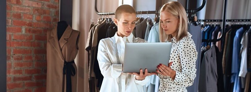 Two young female employees working on social media marketing for fashion brands, standing and holding a laptop in front of a clothing rail and next to a window